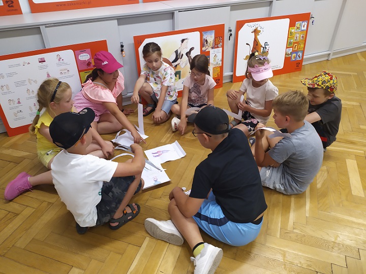 Dzieci siedzące w kręgu na podłodze z papierowych liter układają nazwę stworzenia z rysunku. Za nimi trzy plansze z bohaterami książek Wydawnictwa Zakamarki.