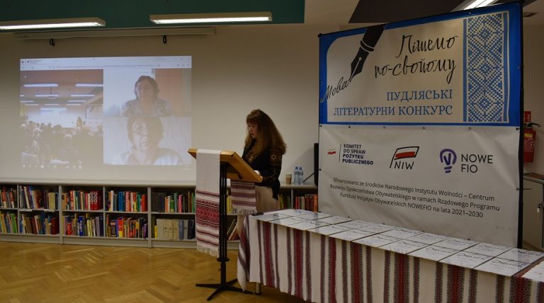 Pośrodku przy pulpicie stoi czytająca kobieta, po lewej stronie ekran z uczestnikami online, po prawej baner z tytułem konkursu "piszemo po swojomu"