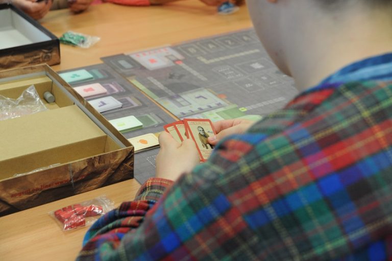 Mężczyzna z kartami w dłoniach tyłem do obiektywu nad planszą gry „Kolejka”