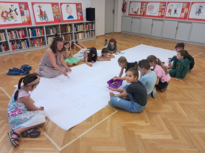 Dzieci rysują na podłodze za nimi półki z książkami. Obok dorosła kobieta