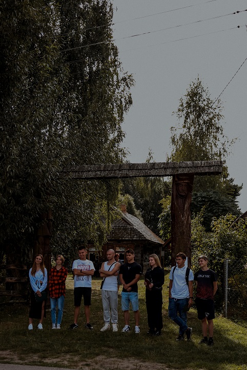 Przed drewnianą bramą do skansenu stoi w rzędzie grupa młodzieży