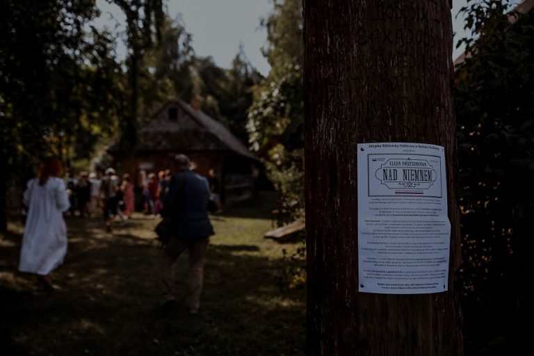 Z prawej strony ogłoszenie o Narodowym Czytaniu na drewnianym słupie, w tle idące w stronę chaty w oddali osoby.