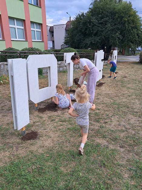Litery w formie przestrzennej, przed czwórka dzieci z dorosłą kobietą. Za fragmenty budynków, drzewo mirabelka