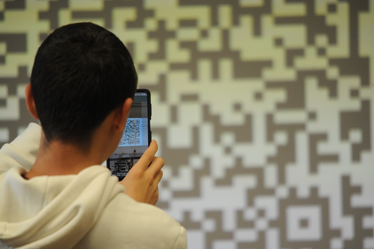 Po lewej tyłem do obiektywu mężczyzna skanujący telefonem kod QR ze slajdu wyświetlonego na ścianie