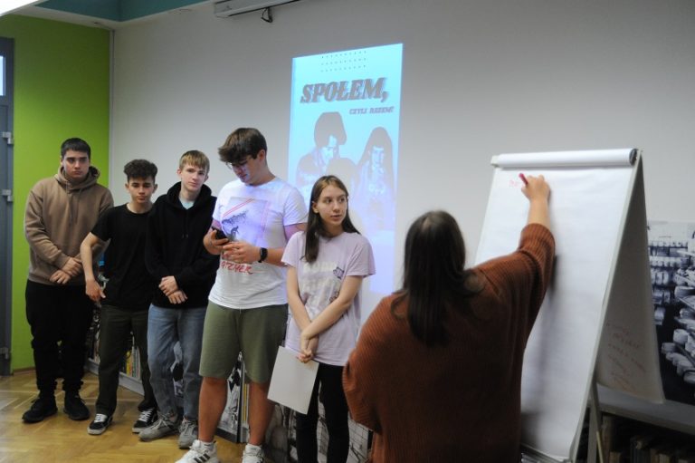 Młoda kobieta oraz czterech mężczyzn stojących w rzędzie przed ścianą z wyświetlonym slajdem z plakatem z napisem „Społem, czyli razem!”. Po prawej tyłem do obiektywu kobieta pisząca na flipcharcie