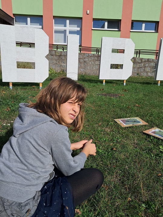 Dziewczynka siedząca po turecku na trawie. Za nią litery BIB. W tle ściana frontowa biblioteki