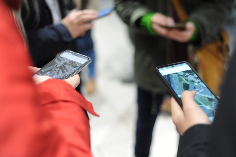 Na pierwszym planie widoczne ekrany dwóch smartfonów z wirtualna mapą w dłoniach dwóch osób. Na drugim – nieostre, ucięte sylwetki dwóch osób ze smartfonami