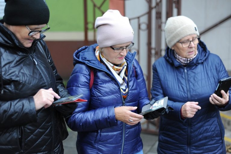 Trzy kobiety w wierzchnich okryciach z telefonami w dłoniach przed wejściem do biblioteki