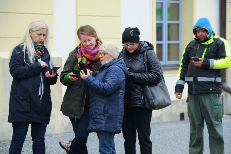 Cztery kobiety oraz mężczyzna w wierzchnich okryciach z telefonami w dłoniach przed budynkiem ratusza podczas udziału w mobilnej grze terenowej „Afera”