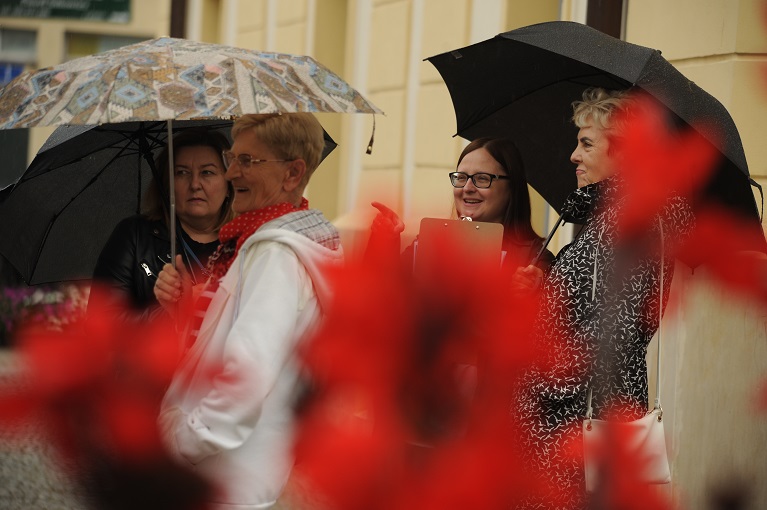 Cztery uśmiechnięte kobiety pod parasolami. W tle ściana ratusza w Bielsku Podlaskim. Na pierwszym planie rozmyte czerwone kwiaty