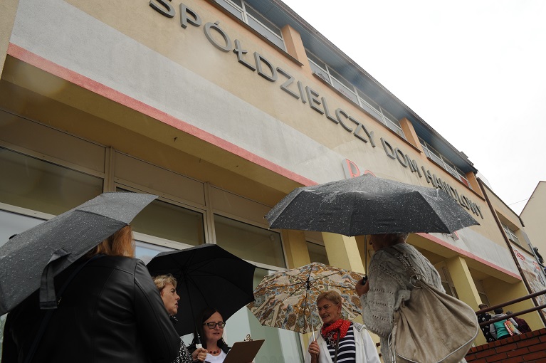 Pięć kobiet pod parasolami przed budynkiem z napisem „Spółdzielczy Dom Handlowy” na elewacji.
