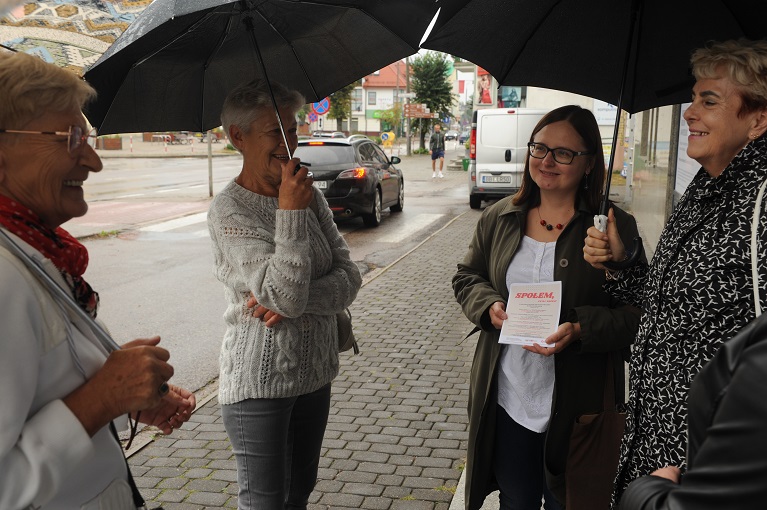 Cztery kobiety pod parasolami na chodniku przed budynkiem dawnego baru „Ratuszowy” przy ulicy Adama Mickiewicza. Kobieta druga z prawej z ulotką z napisem „Społem” w dłoniach. W tle dwa samochody