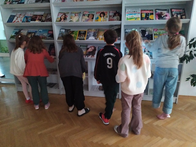 Pięć dziewczynek oraz chłopiec tyłem do obiektywu przed regałem z czasopismami w trakcie poszukiwania listu ukrytego w jednej z gazet