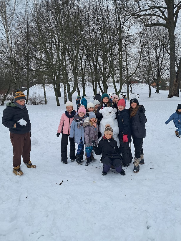 Grupa dzieci ustawiona wokół śniegowego plastusia. W tle drzewa. Za nimi górka. Wszystko pokryte śniegiem