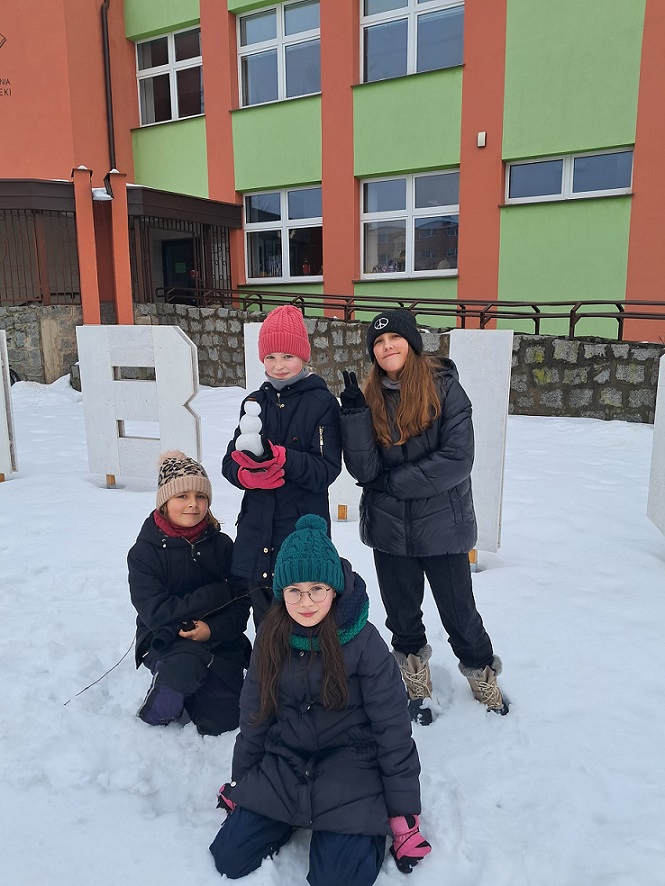 4 dziewczynki ustawione na tle napisu przed budynkiem biblioteki. Jedna z nich trzyma figurkę bałwana ulepioną ze śniegu