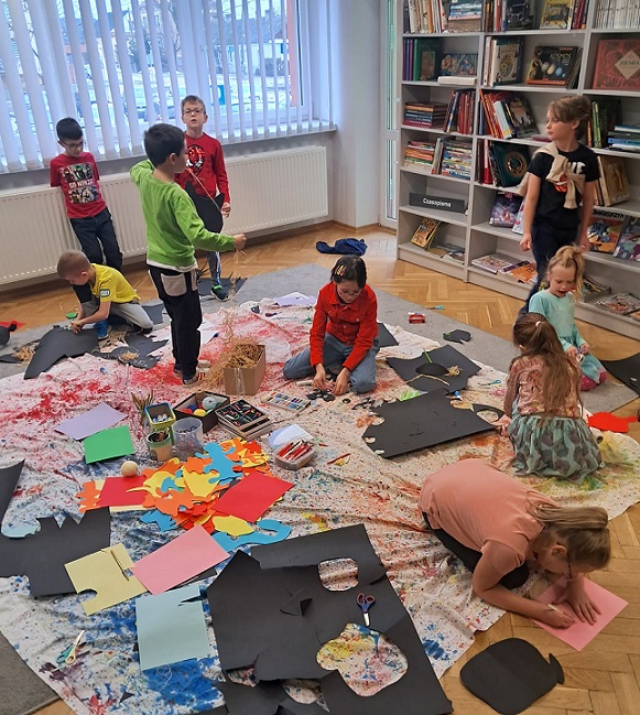 Dziewięcioro dzieci w przestrzeni biblioteki podczas pracy twórczej. Na podłodze kolorowy materiał, papiery