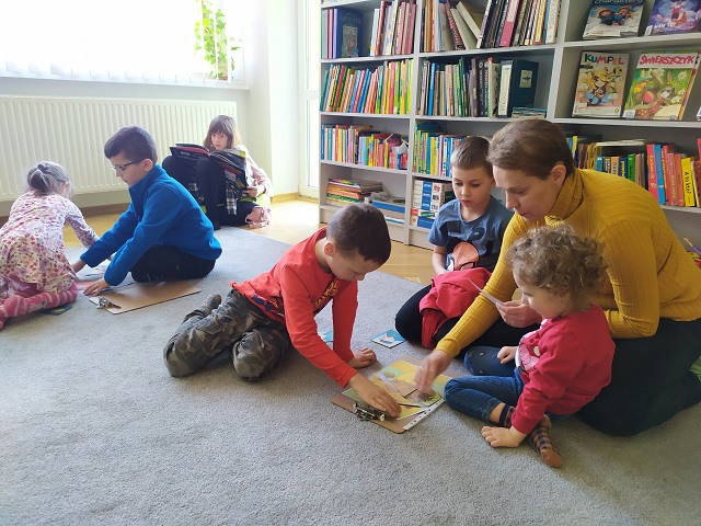 Na dywanie pięcioro dzieci oraz kobieta układają puzzle z kopiami obrazów Vincenta van Gogha z serii ze słonecznikami. Na drugim planie regały z książkami, po lewej dziewczynka czytająca książkę.