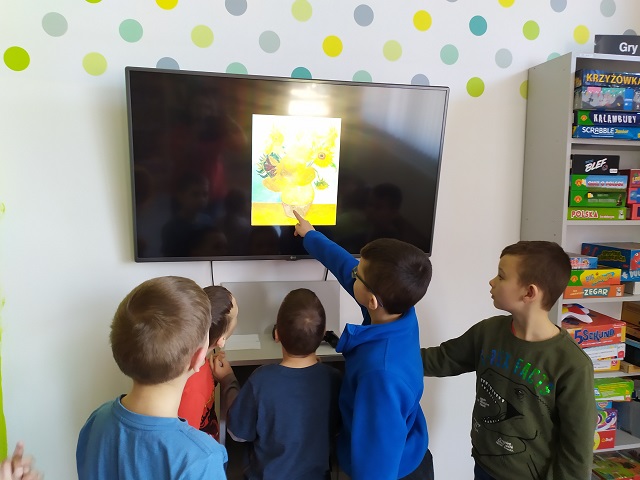 Pięciu chłopców przed ekranem telewizora z wyświetlonym obrazem Vincenta van Gogha „Martwa natura: wazon z dwunastoma słonecznikami”. Jeden z chłopców wskazuje podpis artysty na obrazie.