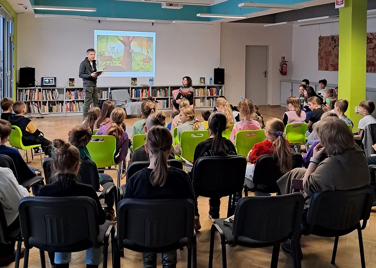 Grupa dzieci i jedna dorosła osoba siedzą w sali bibliotecznej. Przed nimi z lewej strony stojący mężczyzna z książką w dłoniach. Obok kobieta na fotelu. Za nimi wyświetlona na ścianie ilustracja.