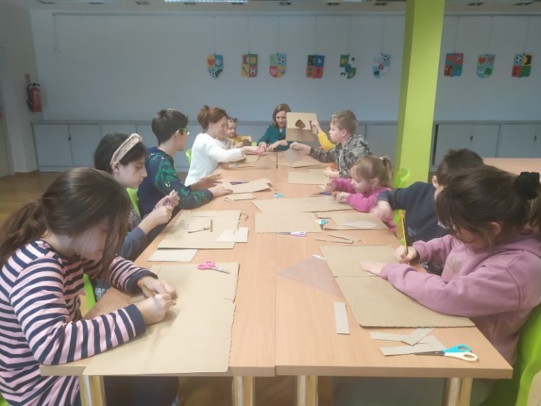 Przy długim stole dzieci i dorośli w trakcie wykonywania pracy plastycznej z kartonu. Na drugim planie na ścianie kolorowe herby z papieru, poniżej szafki