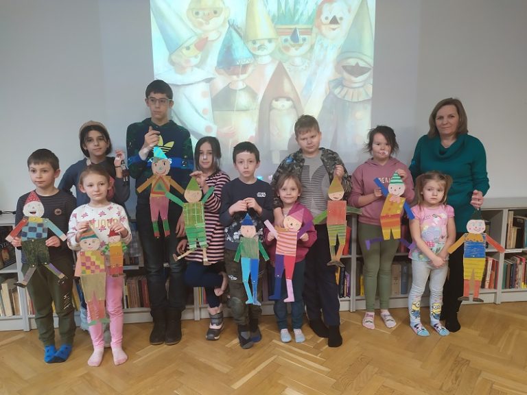 Dziesięcioro dzieci oraz kobieta prezentujący wykonane przez siebie kolorowe pajacyki. W tle slajd z reprodukcją obrazu Tadeusza Makowskiego „Maskarada”. Pod slajdem niskie regały z książkami