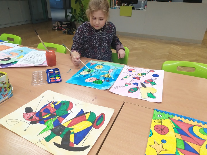 Przy stole dziewczynka podczas uzupełniania szkicu mazakami oraz farbami. Na blacie kolorowe prace inspirowane twórczością Miró