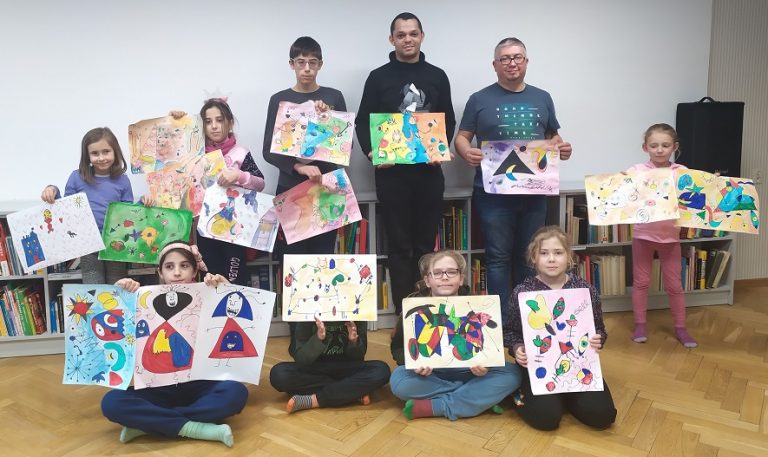 Uczestnicy warsztatów prezentują wykonane przez siebie prace inspirowanej obrazami Miró. Za nimi niskie regały z książkami