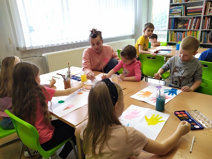 Przy dwóch stołach dzieci i kobieta w trakcie malowania farbami akwarelowymi. W głębi okno oraz regał z książkami