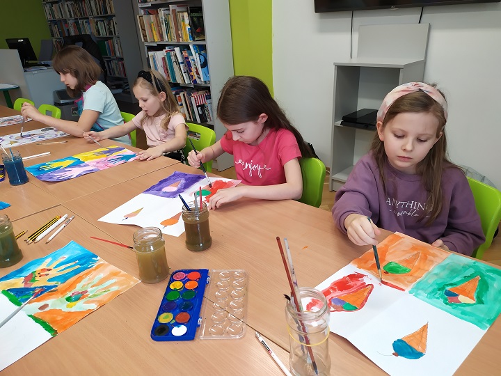 Przy stole cztery dziewczynki w trakcie malowania farbami akwarelowymi. Na drugim planie stanowiska bibliotekarek oraz regały z książkami